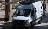 Grey Mercedes-Benz Sprinter Winnebago Revel RV rental, awning extended, parked next to a tree with a folding chair.