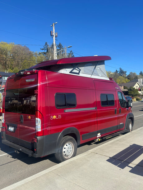 Red Winnebago Solis camper van for RV rental, parked on a sunny street with its pop-top roof extended, showcasing its design.