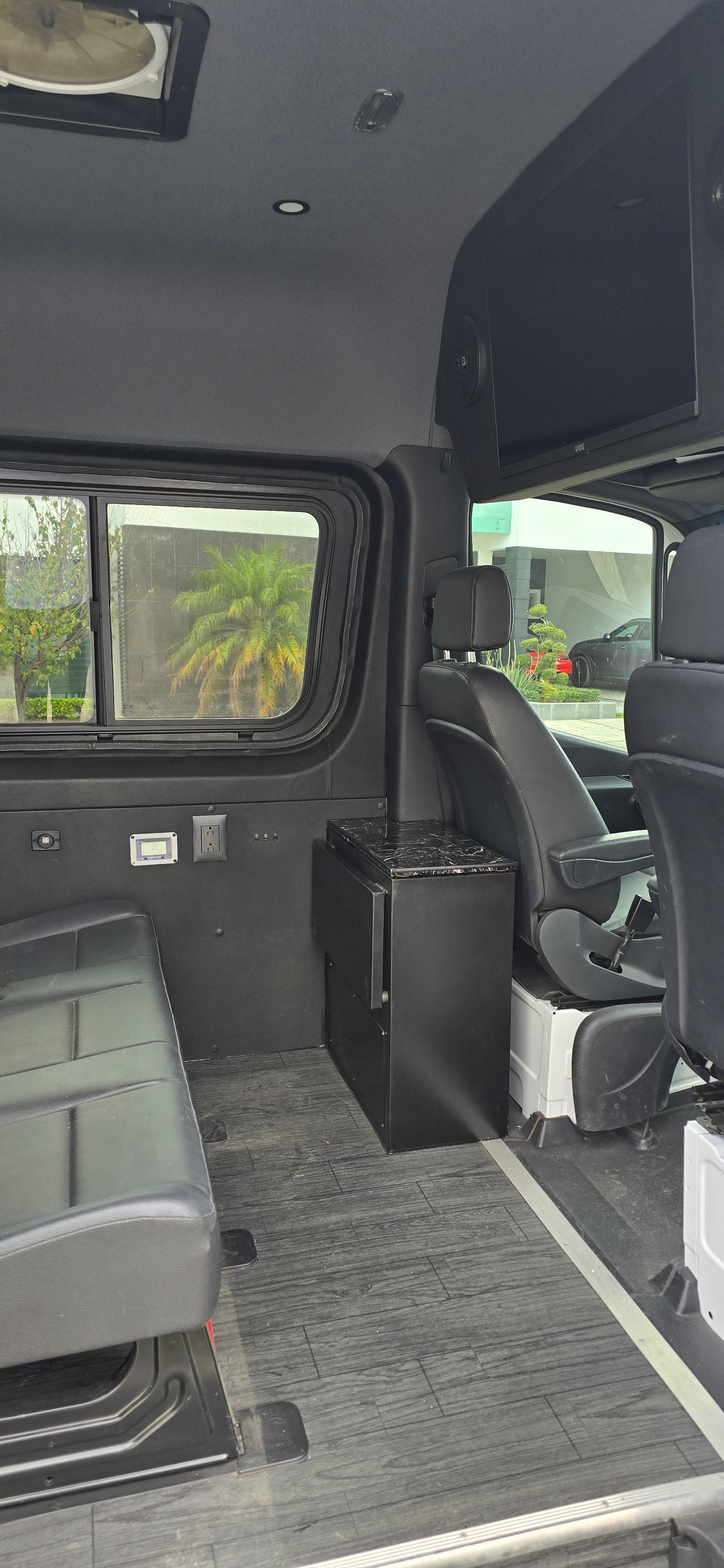 Custom van RV rental interior: black seating, wood floor, TV, outlets, & side window view for comfortable travel/work.