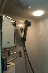 This RV rental's compact bathroom shows a hand-held shower, a multi-angle mirror, and toilet paper, all brightly lit from above.