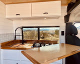 Modern camper van kitchen in an RV rental, with wood counters, stove, sink, and mountain views.