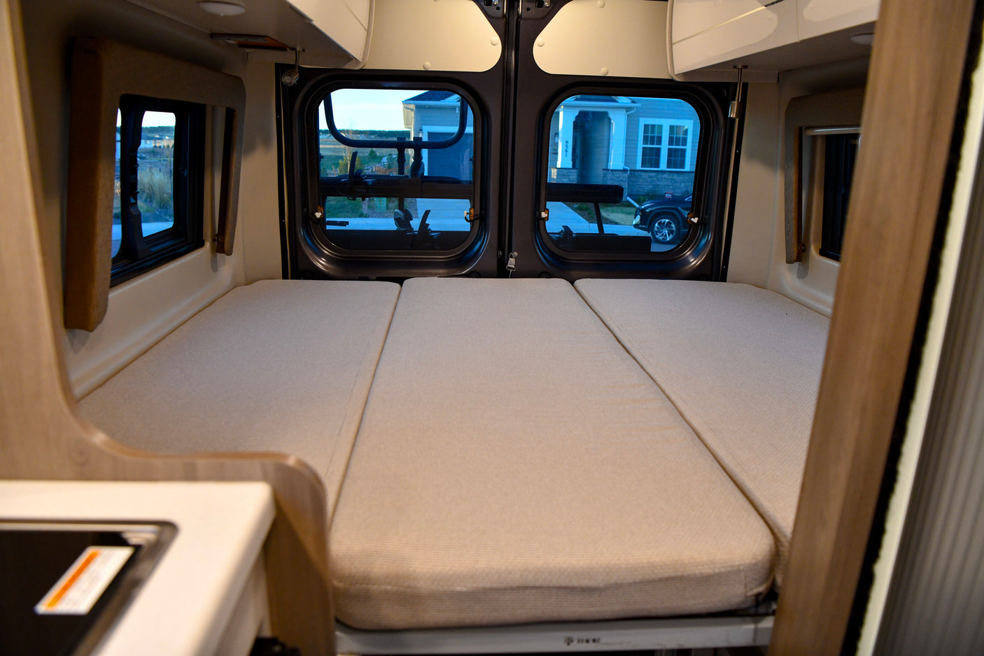 View of a spacious, light-colored bed with three cushions inside an rv rental, featuring rear windows looking out to a home.
