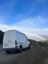 Experience an RV rental adventure! A white Sprinter campervan overlooks a stunning rainbow over hills.