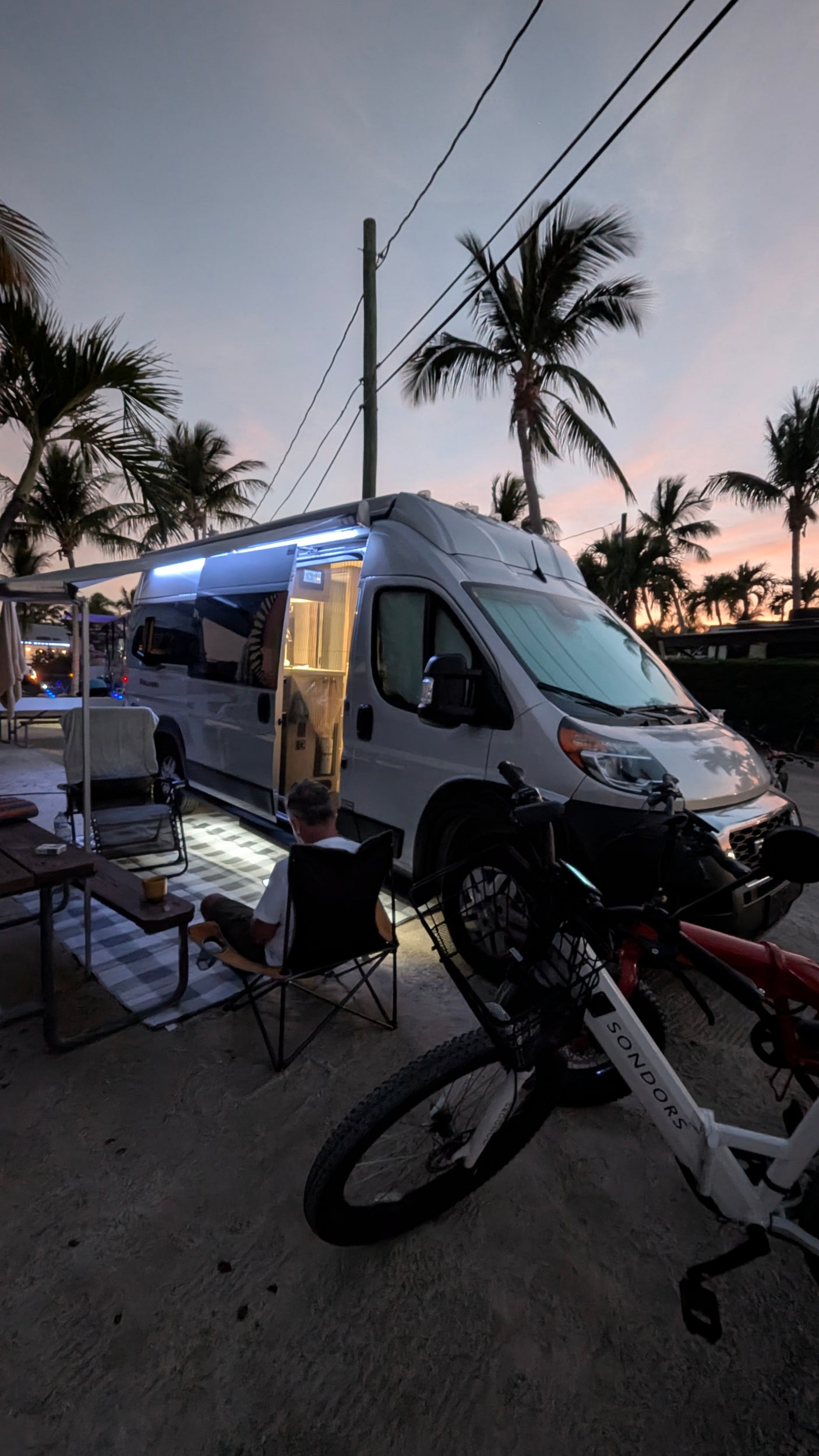 A cozy RV rental camper van scene at dusk, with palm trees, outdoor seating, and Sondors e-bikes. Perfect for travelers.