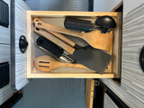 Open RV rental kitchen drawer filled with neatly organized cooking utensils: wooden spoons, spatulas, can opener, peeler.