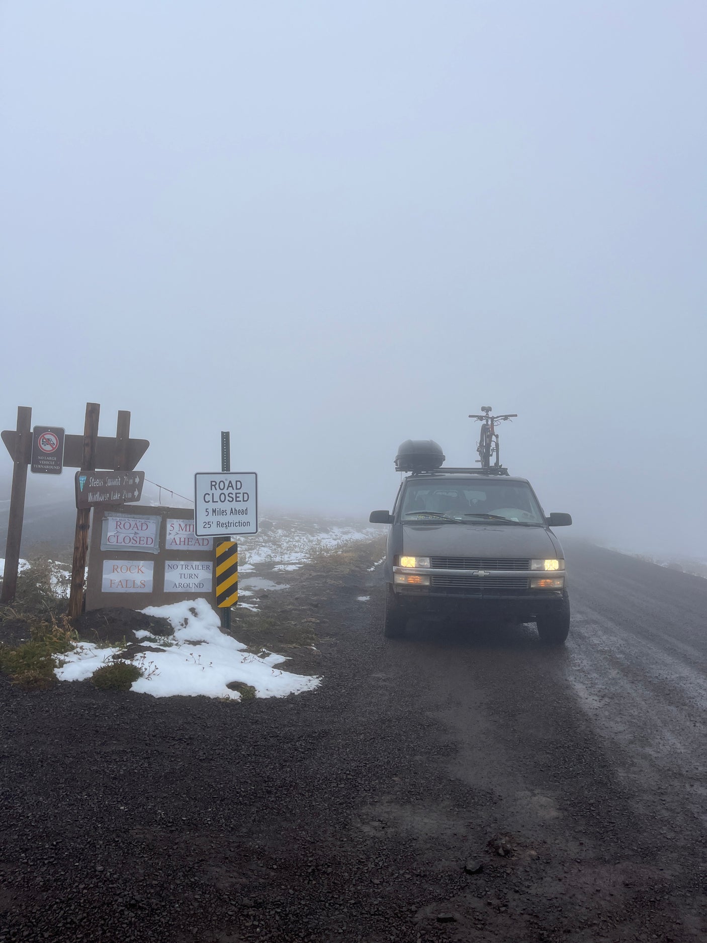 A misty road leads to signs saying 'ROAD CLOSED' for an RV rental van with a bike on top. Snowy terrain at a foggy summit.