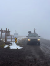 A misty road leads to signs saying 'ROAD CLOSED' for an RV rental van with a bike on top. Snowy terrain at a foggy summit.