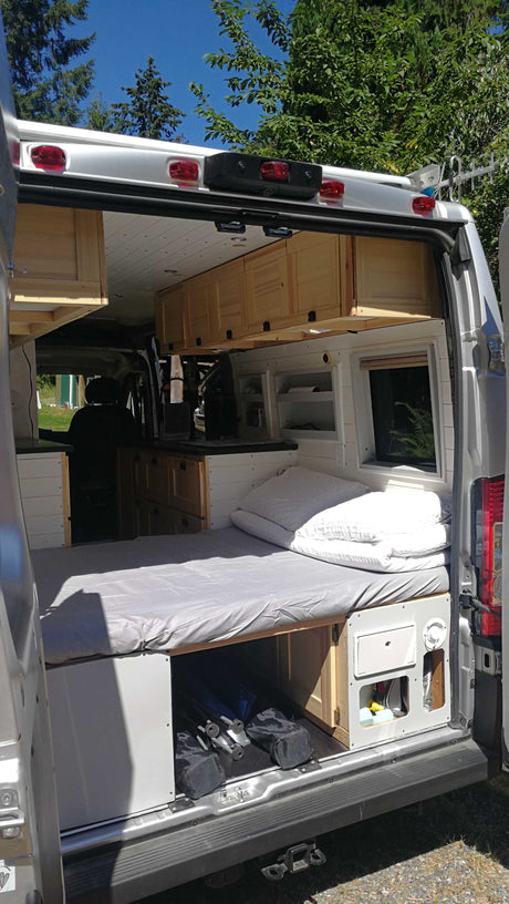 Open back of a custom camper van RV rental. See the comfy bed, kitchen, and wooden cabinets inside.