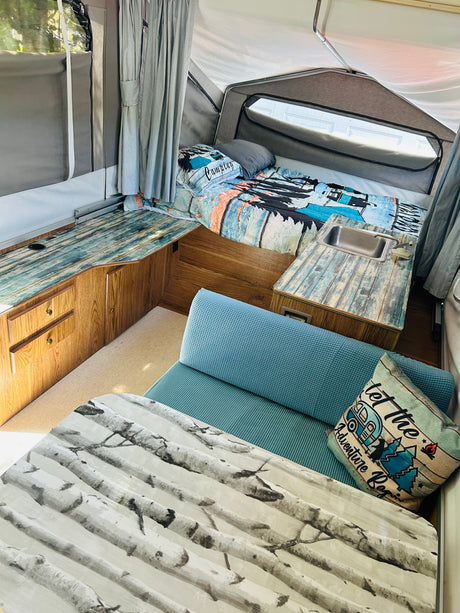 Pop-up RV rental interior: cozy bed, mini kitchen with sink, blue dinette seating, and birch table. Ideal for adventures!