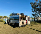 This white camper van, an ideal rv rental, is parked on grass by a waterfront, its doors open to show the interior setup.