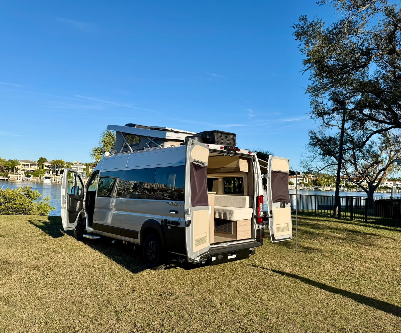 This white camper van, an ideal rv rental, is parked on grass by a waterfront, its doors open to show the interior setup.