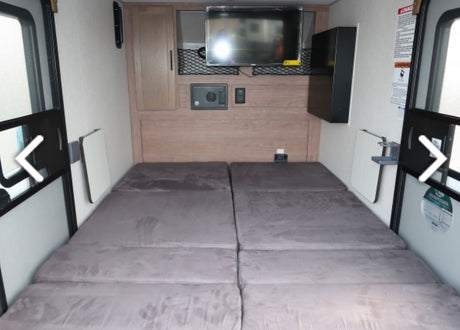 Compact RV rental interior showing two large floor mattresses, a mounted TV, storage cabinet, and foldable side tables.
