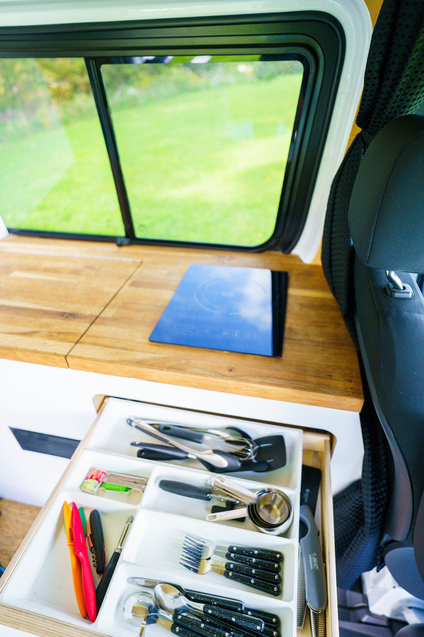 Inside an RV rental, a kitchen space with an organized utensil drawer, induction cooktop, and window view of nature.