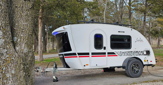 White Luna Rover travel trailer parked at a wooded campsite. Perfect for an RV rental getaway.