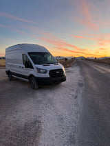 A white Ford Transit camper van (RV rental) parked on a dirt pull-off at sunset, beside a road leading to distant hills.