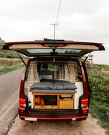Open tailgate of an RV rental camper van showing its cozy bed and interior storage, parked by a road.