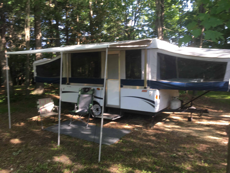 Pop-up rv rental at a serene campsite. A white and blue camper with an awning extends over chairs in the sunny woods. — photo 7