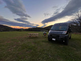 Enjoy a scenic RV rental experience at sunset. This dark van is parked by a cozy fire pit and picnic table in a grassy field.