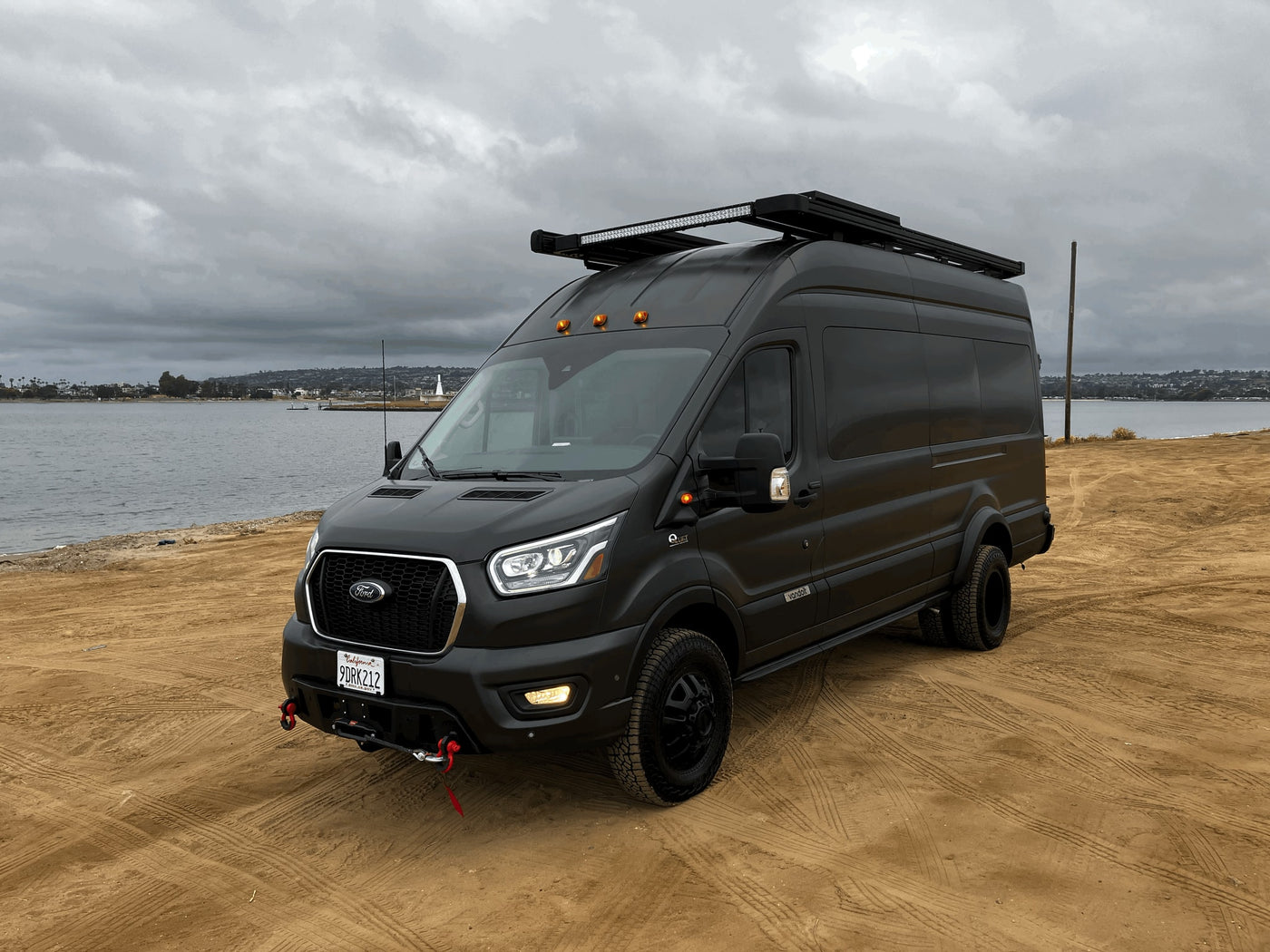 Black Ford Transit adventure van (RV rental) on sand by water. Equipped for off-road journeys with roof rack & rugged tires. — photo 6