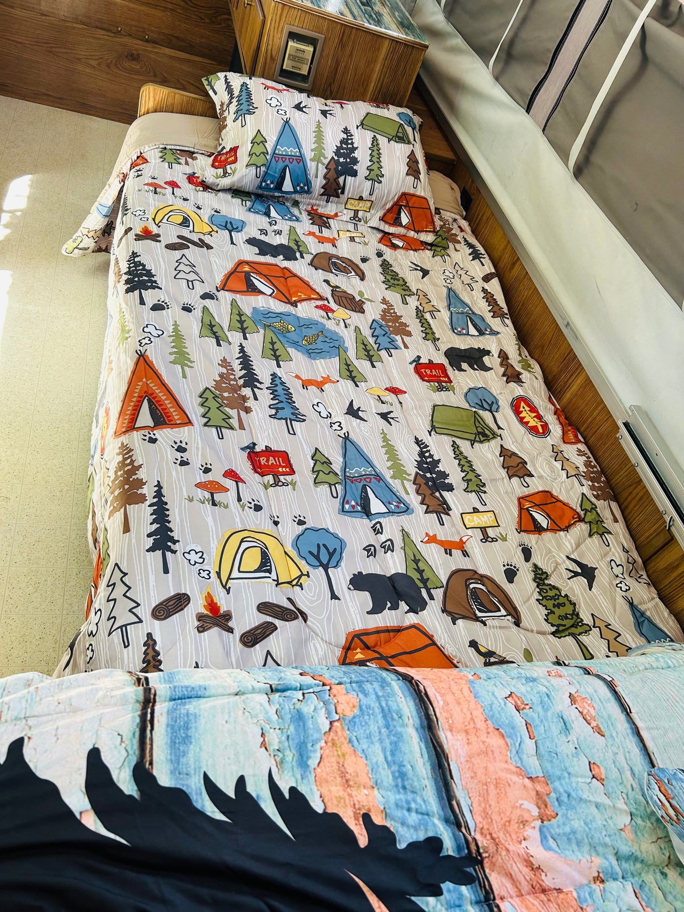 Cozy RV rental bed with camping-themed comforter showing tents, trees, and bears. Ideal for wilderness adventures.