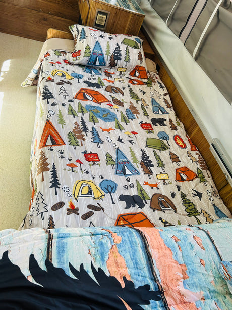 Cozy RV rental bed with camping-themed comforter showing tents, trees, and bears. Ideal for wilderness adventures.