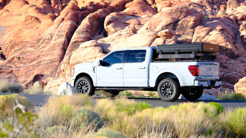 A white Ford F-150 overland truck with a rooftop tent & cargo rack on a desert road, backed by red rock formations. Great rv rental option. — photo 9