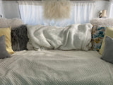 Cozy RV rental bed with soft white blankets and various pillows, including fluffy, shaggy, and floral designs.