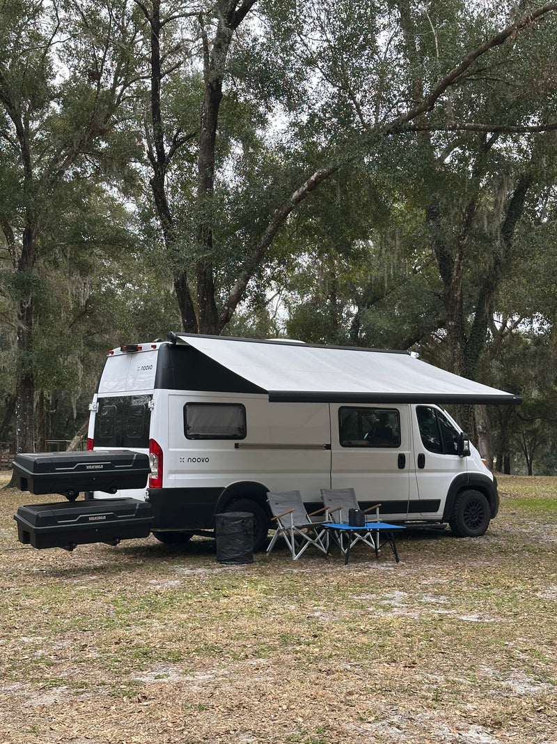 White x noovo rv rental camper van with awning, chairs, and storage boxes. Set up for camping among mossy trees on a grassy outdoor site. — photo 14