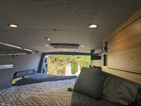 Inside a modern RV rental, a comfy bed with patterned sheets fills the view. Grey walls, overhead lights, & wood cabinets.