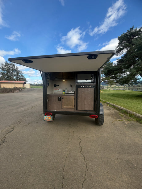 Compact RV rental with an open rear hatch revealing a complete outdoor kitchen setup, including microwave, sink, and fridge.