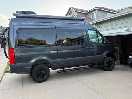 Dark gray Sprinter camper van, ideal for rv rental, parked in a driveway. It features a roof AC, Thule awning, off-road tires & bike rack.