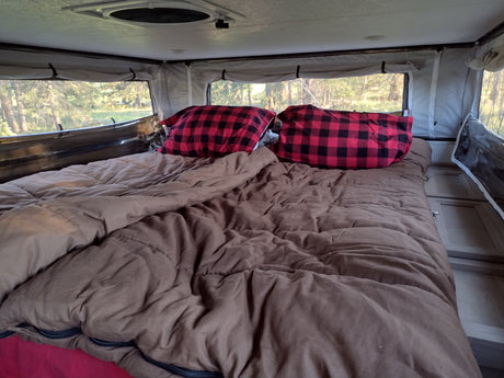 Inside a cozy RV rental: a comfortable bed with red plaid pillows and large windows showcasing forest views.