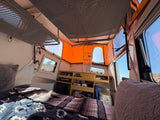 Cozy RV rental interior with bed, storage, and portable toilet. A pop-top roof opens to the sunny outdoors, ready for adventure!
