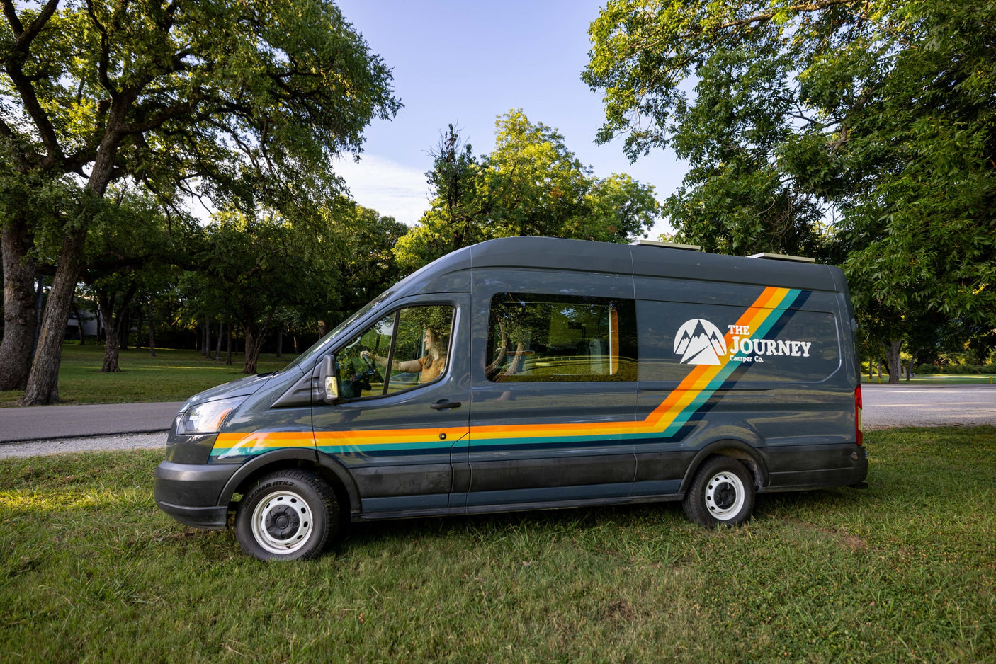  ready for adventure.|A "The Journey Camper Co." dark blue rv rental van parked on green grass with vibrant trees