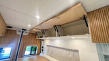 Bright RV rental interior featuring open overhead storage cabinets with gas struts, wood-paneled walls, and built-in lighting.