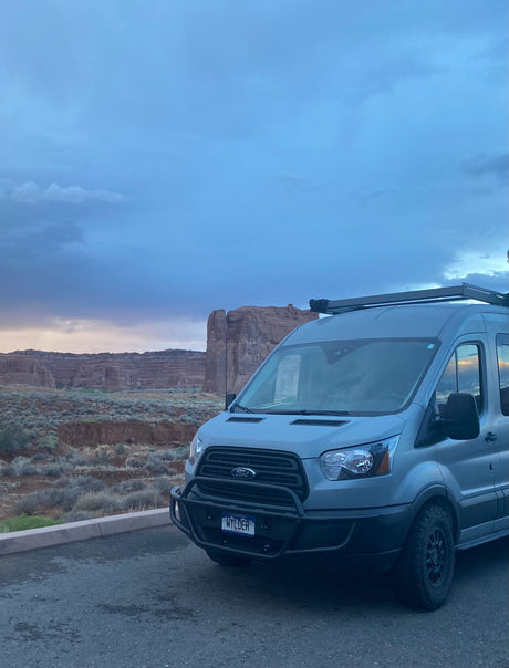 Explore stunning landscapes in this gray Ford Transit RV rental. Parked before majestic rock formations under a dramatic sky.