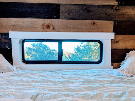 Cozy RV rental bed area with a striped blanket, pillows, and window view of trees. Wood paneling creates a rustic feel.