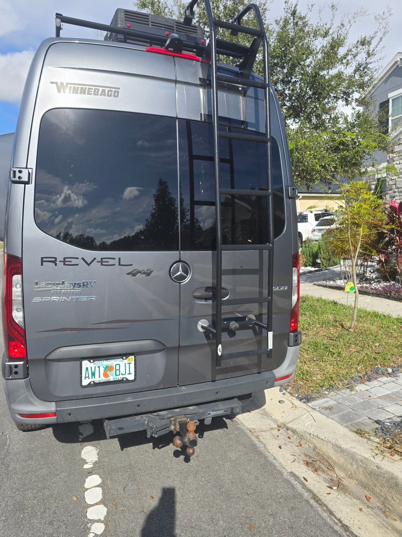 Rear view of a gray Winnebago Revel 4x4 Sprinter RV rental with ladder, tow hitch & Florida plate.