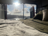 An inviting bed in an rv rental, showcasing a beautiful serene lake and mountain view through the open rear doors on a sunny day.