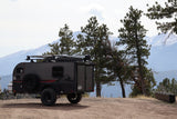 Rugged teardrop RV rental parked on a dirt road, ready for adventure amidst pine trees and snowy mountains.