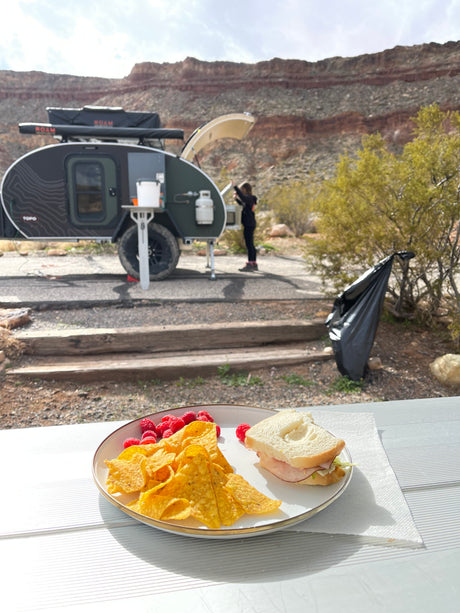 Outdoor meal at a beautiful RV rental campsite. Features a teardrop camper, a person, and scenic desert cliffs.