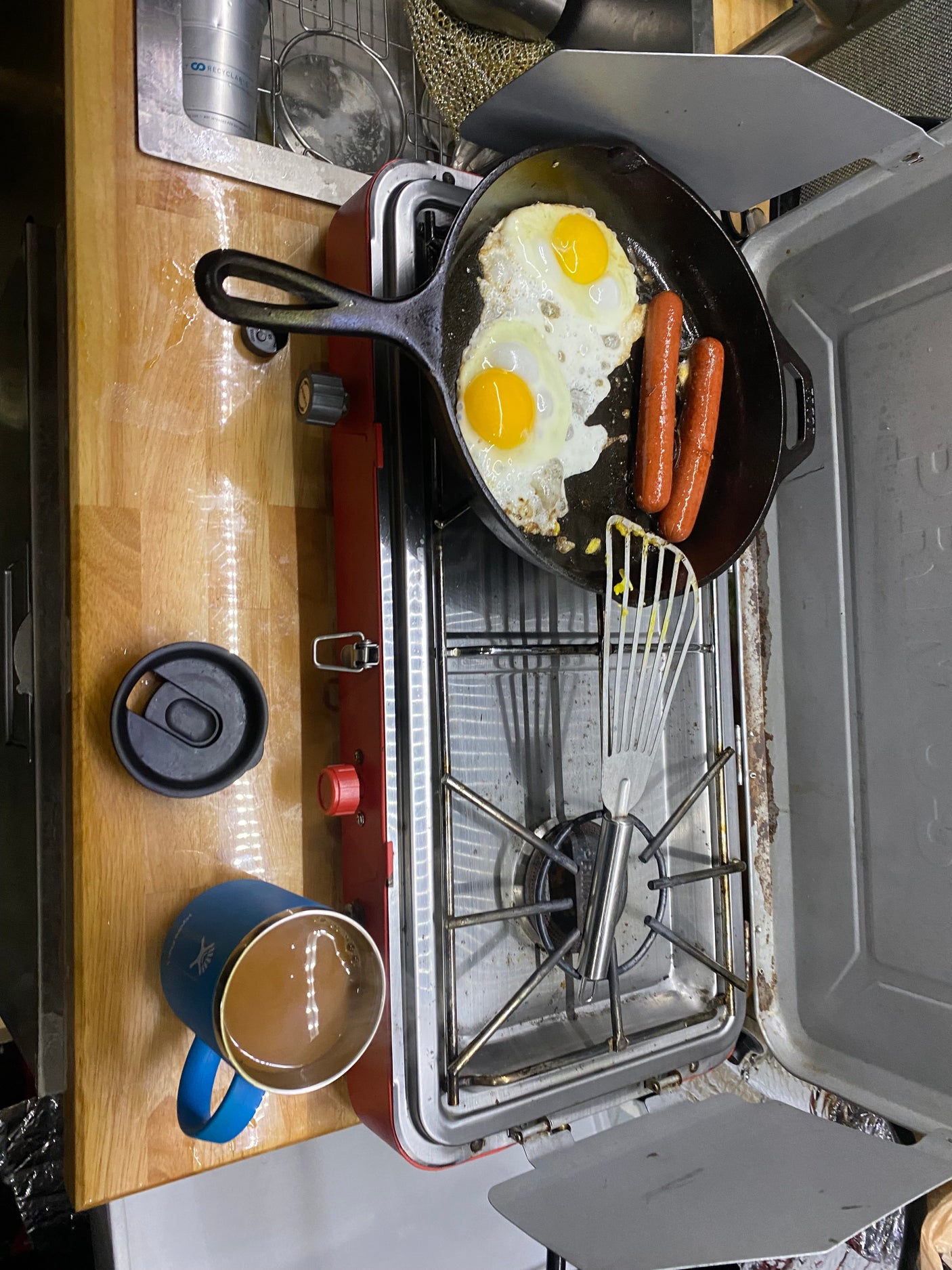 RV rental breakfast: Fried eggs & sausages cooking on a portable stove, with a coffee mug on a wooden counter.