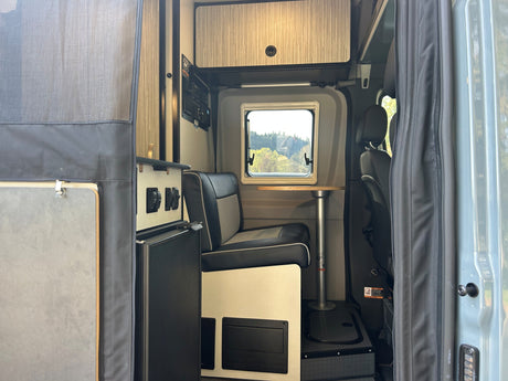 RV rental van interior: compact dining area with bench seating, table, window view, and overhead storage.