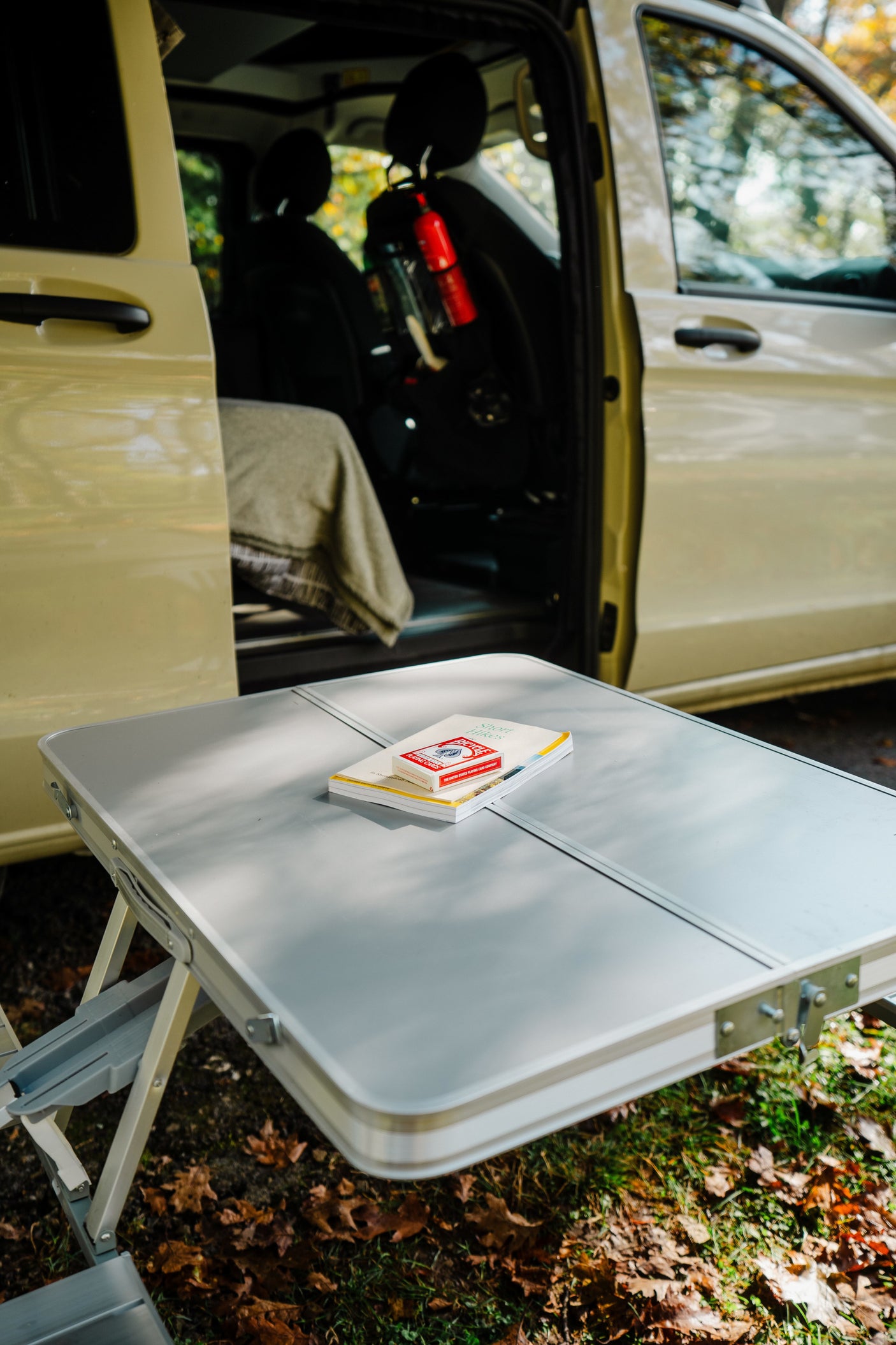 Outdoor relaxation with your RV rental. Silver folding table, book, cards, by a light-colored camper van.