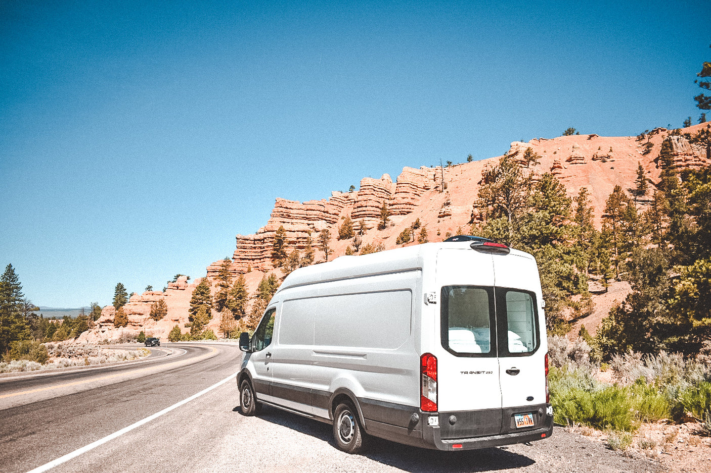 White **rv rental** van parked on a winding road with majestic red rock formations and green pines under a clear blue sky.
