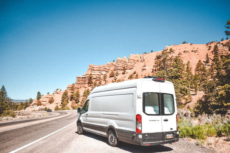 White **rv rental** van parked on a winding road with majestic red rock formations and green pines under a clear blue sky.