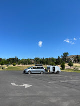Silver minivan with rooftop cargo box and an attached teardrop RV rental trailer, parked in a sunny outdoor lot.