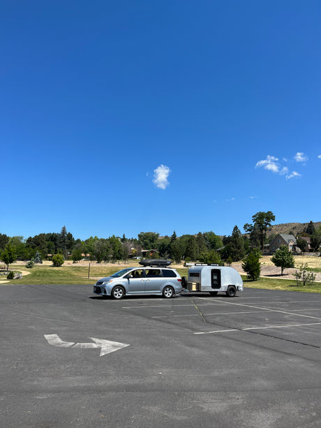 Silver minivan with rooftop cargo box and an attached teardrop RV rental trailer, parked in a sunny outdoor lot.