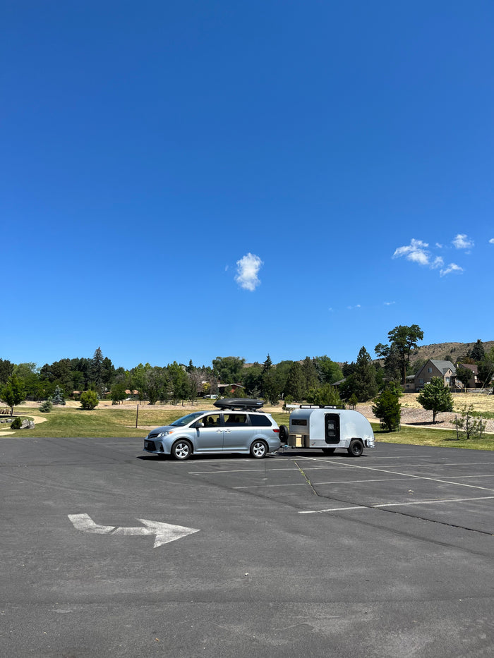 Silver minivan with rooftop cargo box and an attached teardrop RV rental trailer, parked in a sunny outdoor lot.