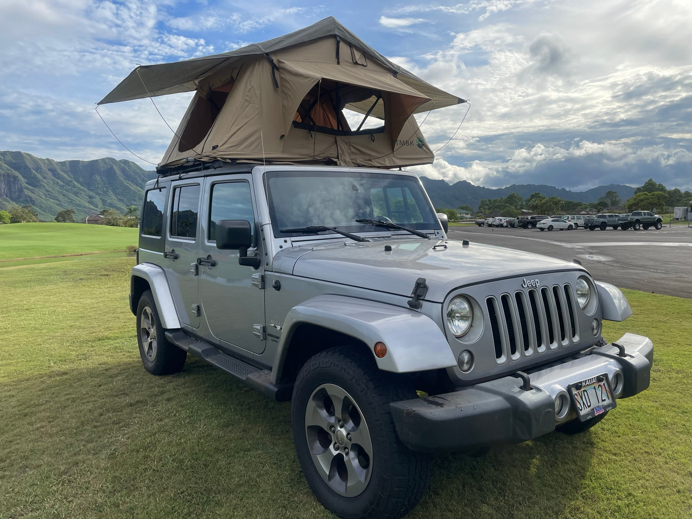 Silver Jeep Wrangler with a rooftop tent on green grass in Kauai. An ideal rv rental for outdoor adventures near mountains.